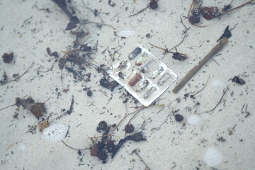 plastic waste and debris, including a blister pack of pills and a plastic fork, floating in shallow seawater among small twigs and leaves — highlighting marine pollution and human impact on coastal 