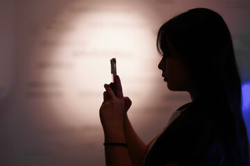Silhouette of a black-haired young girl holding a smartphone in her hand, looking forward into the room.