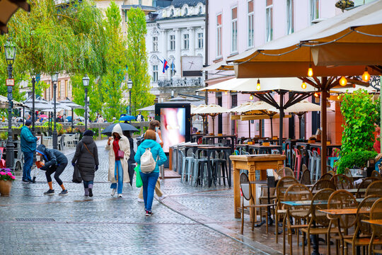 Pedestrians walk past sidewalk cafes and shops along the Ljubljanica River at the riverfront corso promenade after an autumn rain in the picturesque old town of Ljubljana, Slovenia.
