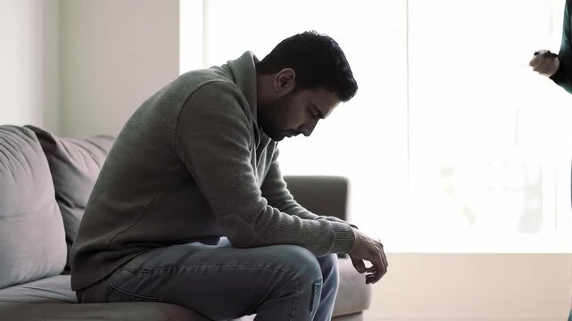 A man sits dejectedly on a couch, head down, hands clasped. A woman in the background