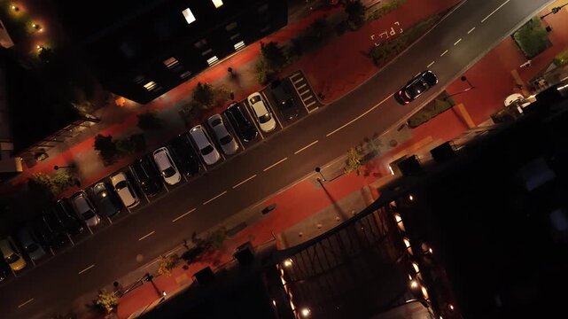 Aerial night shot of a quiet residential street in Paupys, Vilnius, showing parked cars, illuminated sidewalks, modern buildings, and organized urban layout.