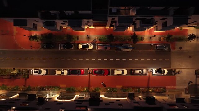 Aerial night shot of a quiet residential street in Paupys, Vilnius, showing parked cars, illuminated sidewalks, modern buildings, and organized urban layout.