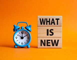 A vibrant orange background highlights a blue alarm clock beside stacked wooden blocks that spell "WHAT IS NEW," implying a sense of anticipation