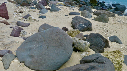 A rocky tropical beach with large stones scattered on golden sand near the ocean. The scene captures raw coastal beauty and natural textures untouched by humans.
