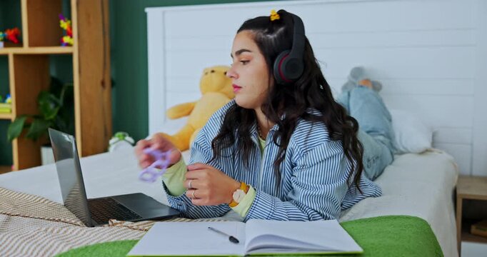 Student, laptop and woman writing notes in home with fidget toy for adhd or audio in bedroom. Music, headphones and girl in bed for learning, education and study on book for exam with sensory tangle