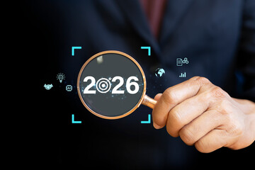 Businessman holding magnifying glass focusing on year 2026 with digital icons of innovation, AI,...