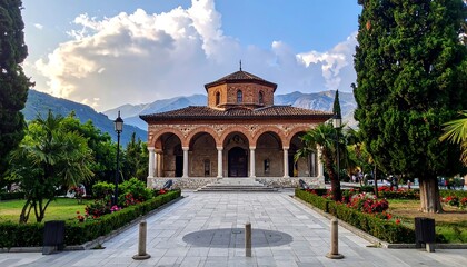 Obraz premium A stone building with arched entrance sits at the end of a stone path, framed by trees and mountains under a cloudy sky