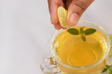 A woman hand squeeze lemon in the green tea. Ginger, half a lemon, honey and basil leaves in a bowl on a burlap top view. Useful vitamin ingredients in a cup of tea on a white background.