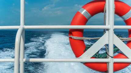 a bright orange life preserver ring is secured to the ship's white metal railing, set against the backdrop of the deep blue ocean and cloudy sky. the boat's wake is clearly visible.
