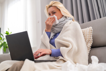 Blonde middle aged woman sitting on a sofa in a cozy room feeling unwell but continuing remote...