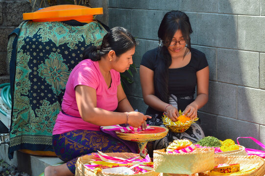 A mother finishes weaving colorful offerings while her daughter carefully places gemitir flowers atop a white and yellow canang sari arranged on a carved wooden bowl.