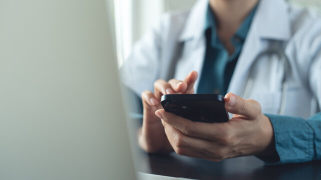 Doctor using smartphone at doctor's office, messaging with patient, prescribing treatment with laptop computer on table at medical clinic, closeup, health care and medicine, consultation online