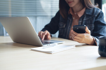 Asian business woman using smartphone during remote working on laptop computer at modern office. Woman using mobile phone surfing the internet, remote working on laptop at coffee shop