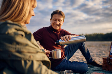 Happy mature man pouring his wife cup of tea on picnic in nature in autumn.