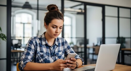 Young woman using mobile phone while working on laptop in modern office