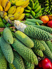 Ripening Monstera deliciosa fruits are tightly stacked on a market counter. Bunches of yellow and green bananas, as well as other vibrant tropical fruits, are visible in the background.