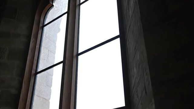 Gothic style arched window in a dark stone wall illuminated by brilliant white light from outside offering a striking architectural contrast