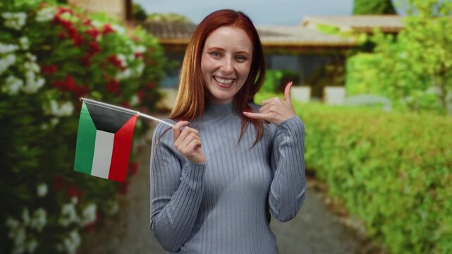 Woman posing outdoors with kuwait flag making call gesture bright smile lush background city street setting vibrant atmosphere.