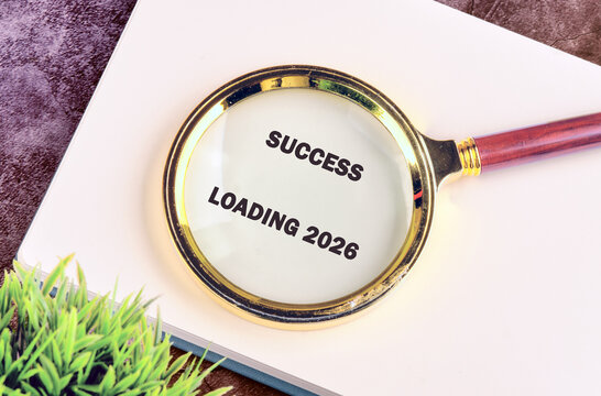 A magnifying glass rests over a plain surface, displaying the phrase success loading 2026. The scene conveys inspiration and motivation toward future achievements and aspirations