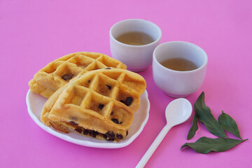 Chocolate chip waffles served on a white plate with two cups of green tea on a pink background. Cozy breakfast setup for tea time, bakery advertisement, or relaxing morning food concept.