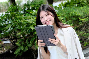 Fototapeta premium 屋外の緑に囲まれ笑顔を見せる、タブレットPCを持つアジア人女性のポートレート。リモートワーク、デジタルライフ、ビジネスとリラックス、テクノロジー、環境調和を表現するイメージ素材、フレッシュな雰囲気で採用広告にも最適