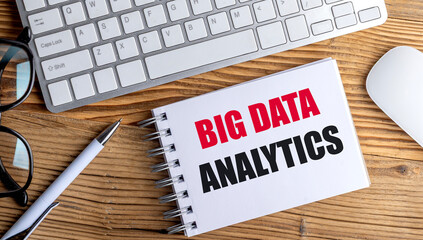Big data analytics text on spiral notepad with keyboard, mouse, glasses and pen on wooden desk