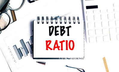 Debt ratio text on spiral notepad with charts, glasses, calculator and pen on white office desk