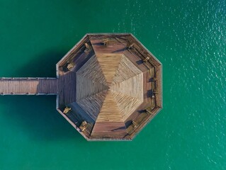 Aerial view of a wooden octagonal pavilion extending over vibrant turquoise water, connected by a narrow walkway.