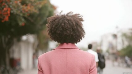 Smiling woman with short curly hair in a pink coat and white shirt, standing on a city street.