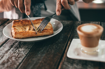 Close-up of a person cutting a slice of apple pastry with a fork and knife on a wooden table, next to a cup of coffee. Cozy breakfast or snack moment in a café setting