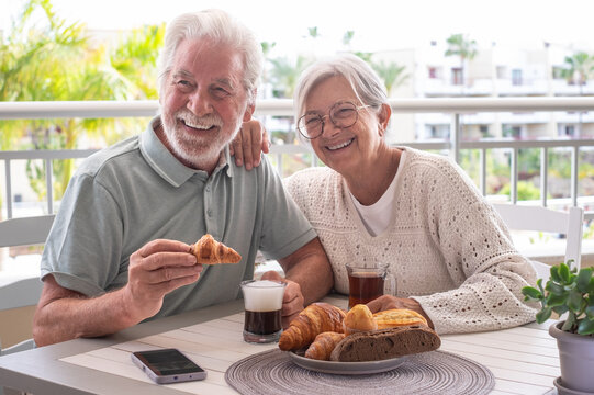 Cheerful senior retired couple enjoying breakfast together sitting outside on home terrace with coffee, tea and baked goods. Peaceful retirement lifestyle.