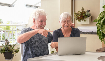 Happy senior couple sitting together at home using a laptop on terrace. Elderly man and woman enjoying time online, browsing the internet. Concept of technology, communication, retirement lifestyle