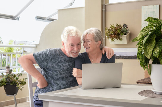 Happy senior couple sitting together at home using a laptop on terrace. Elderly man and woman enjoying time online, browsing the internet. Concept of technology, communication, retirement lifestyle
