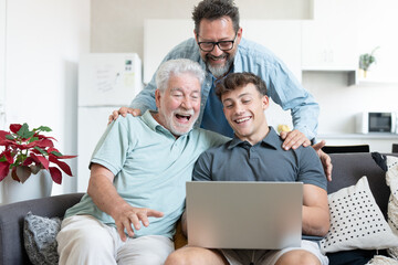 Three generations of men, grandfather, father, and son sitting together on sofa, laughing and enjoying time while using laptop at home, symbolizing family bonding and connection across generations