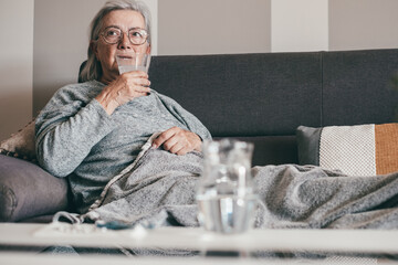 Senior Caucasian woman sitting on the couch with a blanket showing cold and fever symptoms like seasonal flu or Covid takes an effervescent tablet