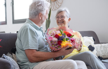 Senior man giving bouquet of flowers at his wife sitting on the sofa at home for anniversary or San Valentines’ day. Pensioners enjoying surprise together. In love people having fun.