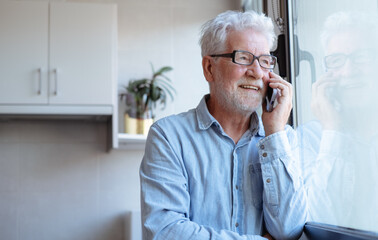 Smiling elderly bearded white haired senior man with eyeglasses looking out of window while talking on mobile phone.