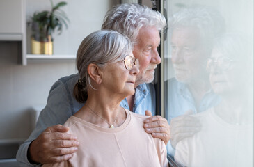 Pensive elderly mature senior couple embracing looking in distance out of window, thinking of personal problems. Old bearded male hugging sad wife close to him, copy space