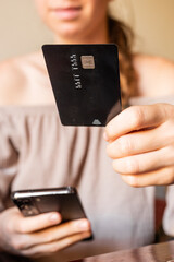 A young Caucasian woman holds a smartphone in one hand and a credit card in the other. She is engaged in online shopping or payment activity.