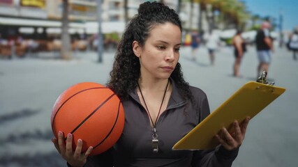 Woman holding basketball and clipboard outdoors on a busy street, focused, wearing sports attire, with blurred people in the background, embodying sports leadership. - Powered by Adobe