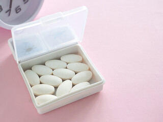 The white pill box is open and contains white plastic pill organizer and clock on pink table. Health, wellness, and care pharmacology concept. Close up.