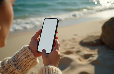 Woman hands hold modern smartphone with blank white screen mockup. Ocean waves, golden sand, and rock are in the background. Traveler wears a cozy knitted sweater. Person uses tech at calm sea shore.