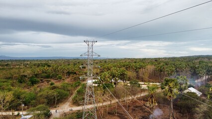 The shot features a high-voltage electrical transmission tower and its thick power cables running through a rural area, with farmland and a winding dirt road visible below