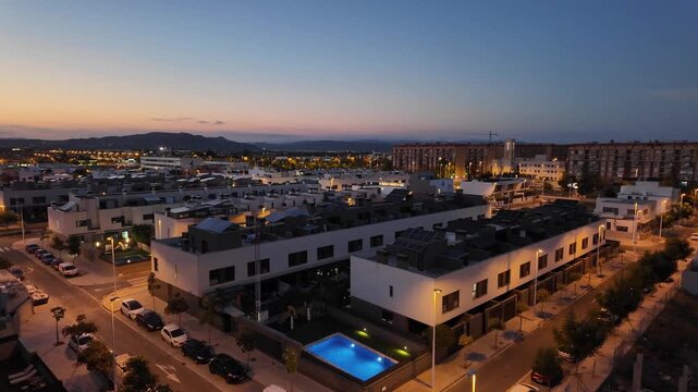 A city skyline at sunset. Lights turn on in homes as the sky changes colors. A pool is visible in the foreground with a quiet street lined with cars.