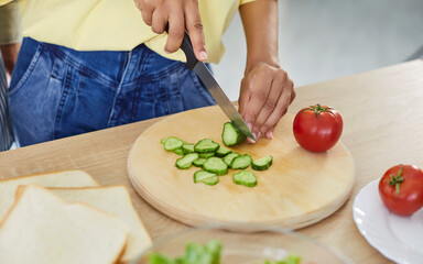 Hands cutting fresh vegetables for salad, slicing cucumber on round wooden board in kitchen. Woman holding knife to cut slices for sandwich preparation, cook healthy diet meal for lunch or dinner