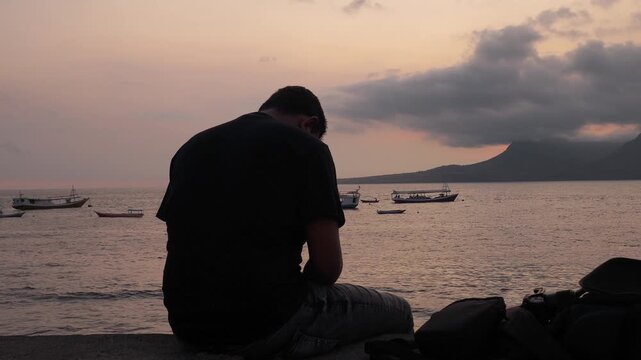 A serene video of a man sitting alone on a seashore at dusk. He is watching the fishing boats on the calm sea, with a mountain island in the background. A peaceful and contemplative scene.
