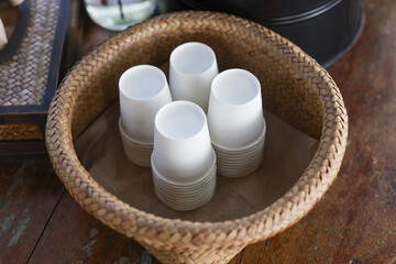 Four white paper disposable cup stand ready within round woven rattan basket. Neat arrangement provides refreshment service, creating an inviting and prepared atmosphere for guests