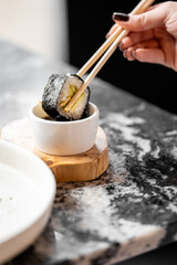 Close-up of a hand with dark nail polish holding sushi with chopsticks, dipping into soy sauce. Minimalist Japanese dining scene on marble surface with wooden coaster.