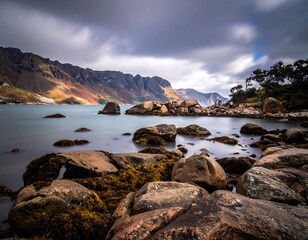 Long exposure seascape showing calm, teal water, rocky shoreline and mountains under a dramatic sky