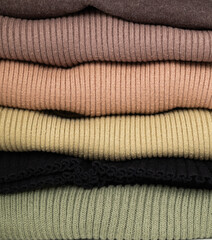 A close-up stack of neatly folded knitted ribbed sweaters in a neutral, earthy color palette: brown, beige, olive, black, and sage green.